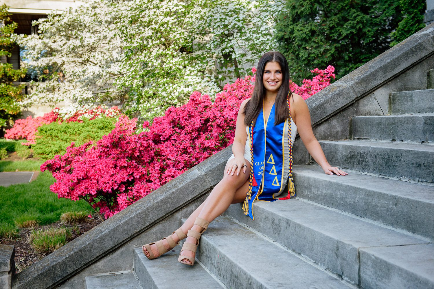 Female College Graduate sits on steps in front of pink flowers