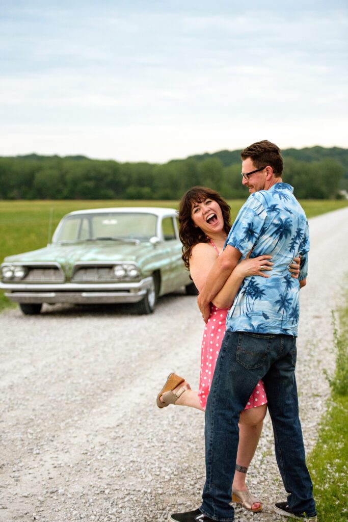 Couple playfully hug in front of vintage car on gravel