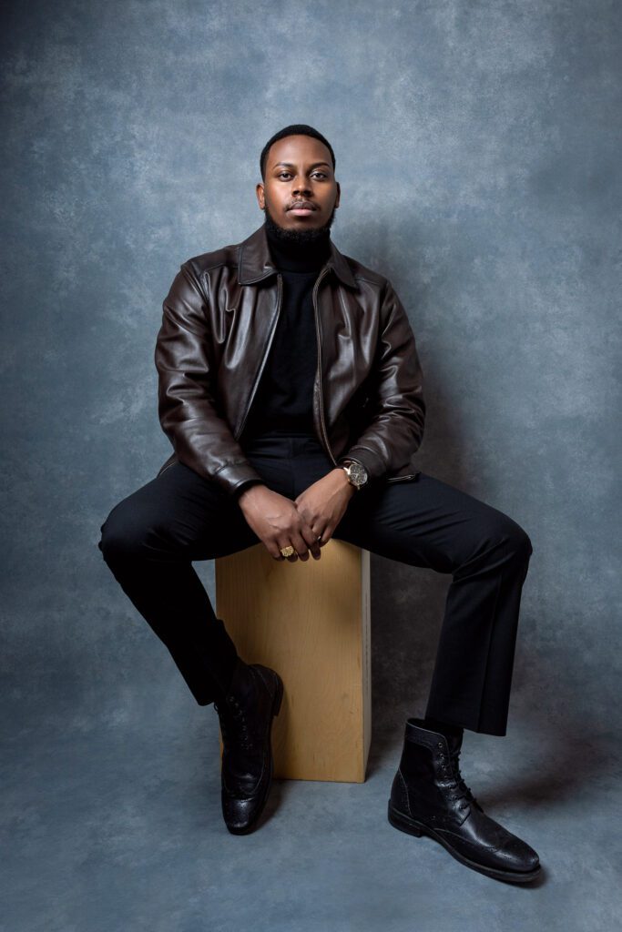 Black male model wearing leather jacket and black pants sitting on apple box