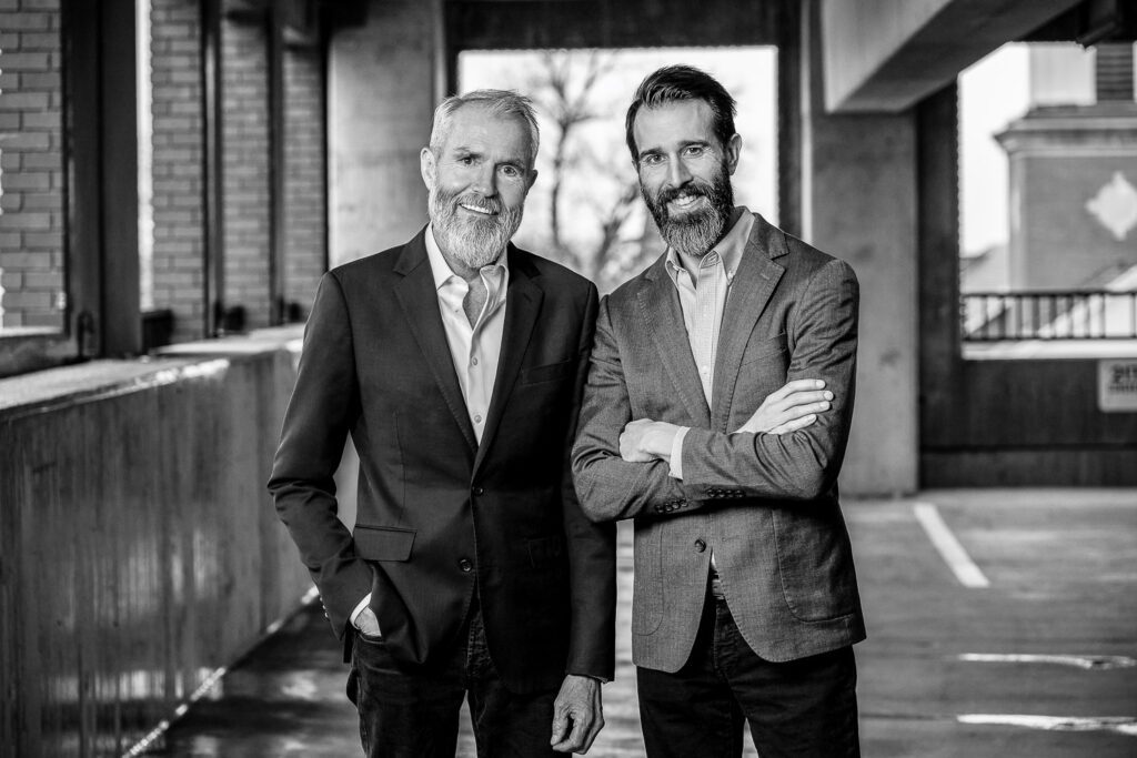 Two men wearing suit jackets pose for portrait in parking garage.