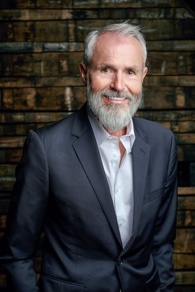 Older man with gray hair and beard poses for a headshot on a wall made out of whiskey barrel staves.
