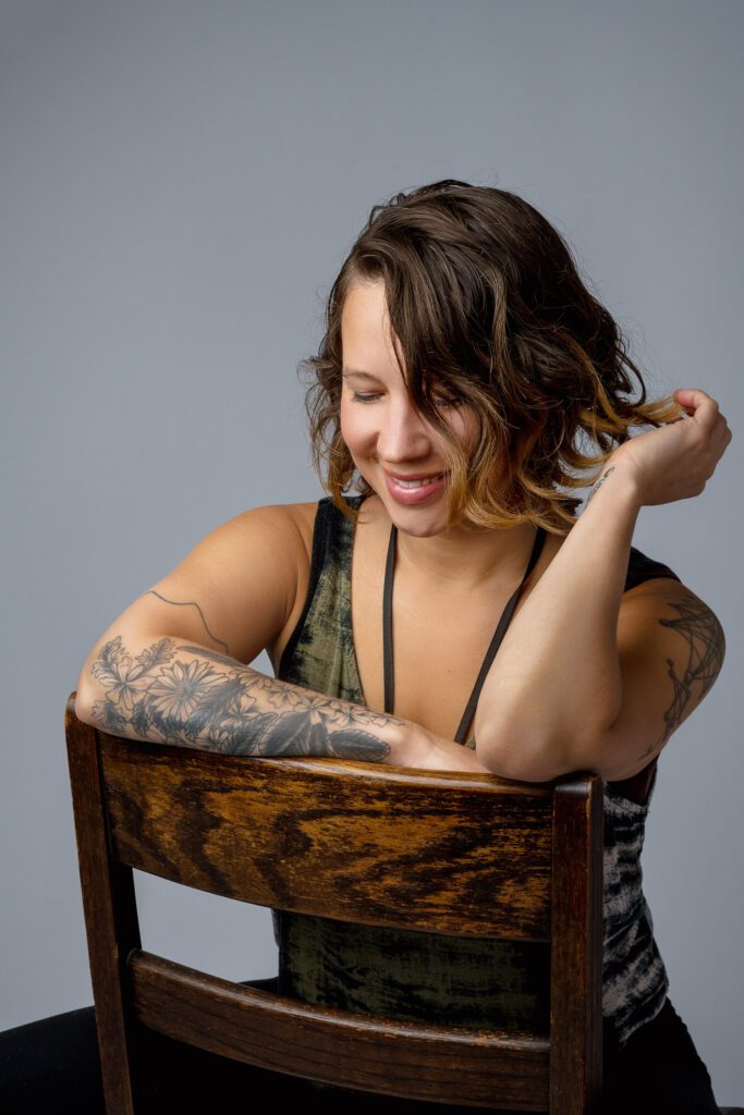 Woman with tattoos and brown hair looks down while sitting backwards on chair.