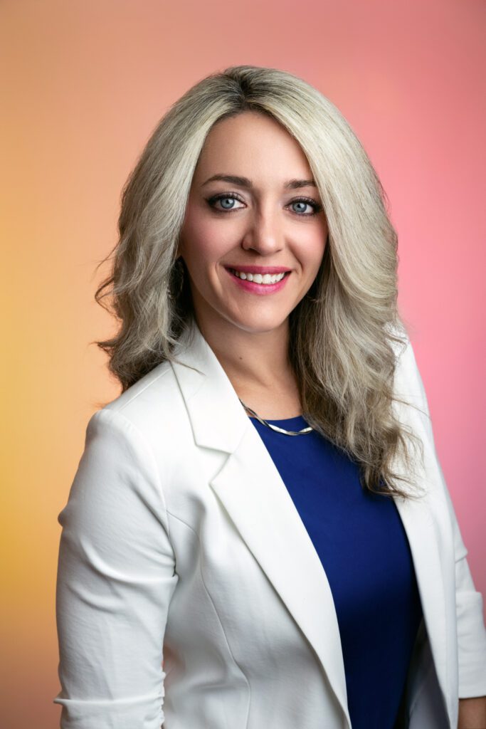 Blonde woman wearing white blazer and blue shirt smiles with orange and pink background
