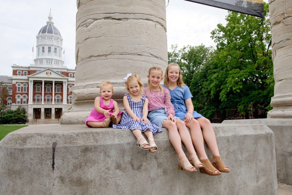 Four sisters sit next to each other on a Mizzou Column in front of Jesse Hall.