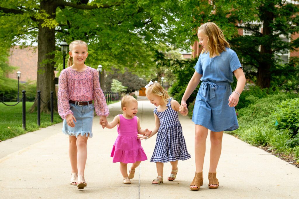 Four sisters holding hands walking down sidewalk during Mizzou Campus Family Photos.