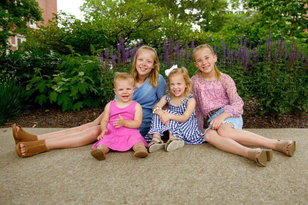 Sisters wearing blue and pink sit in front of purple flowers.