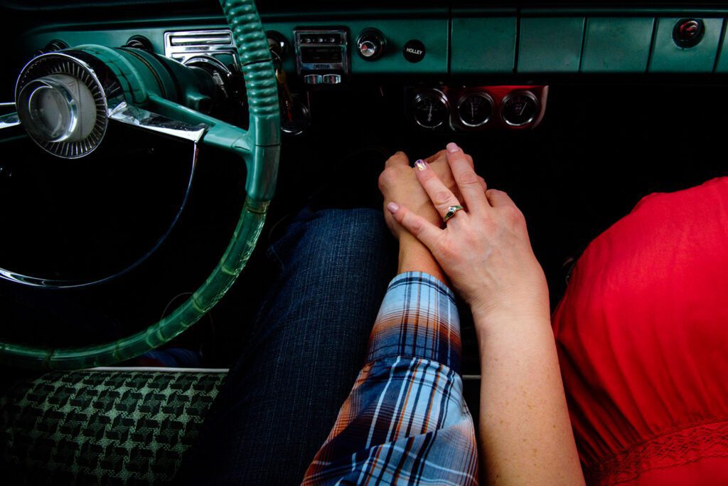 Couple holds hands on gear shift wearing engagement ring.