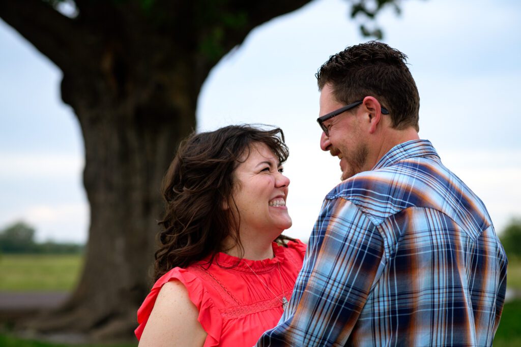 Couple smile at each other under tree.
