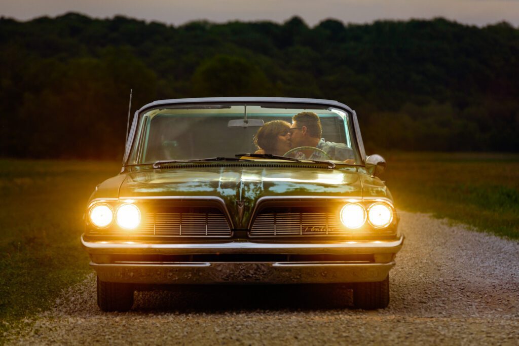 Couple kiss in cab of Pontiac Catalina with headlights on at dusk.