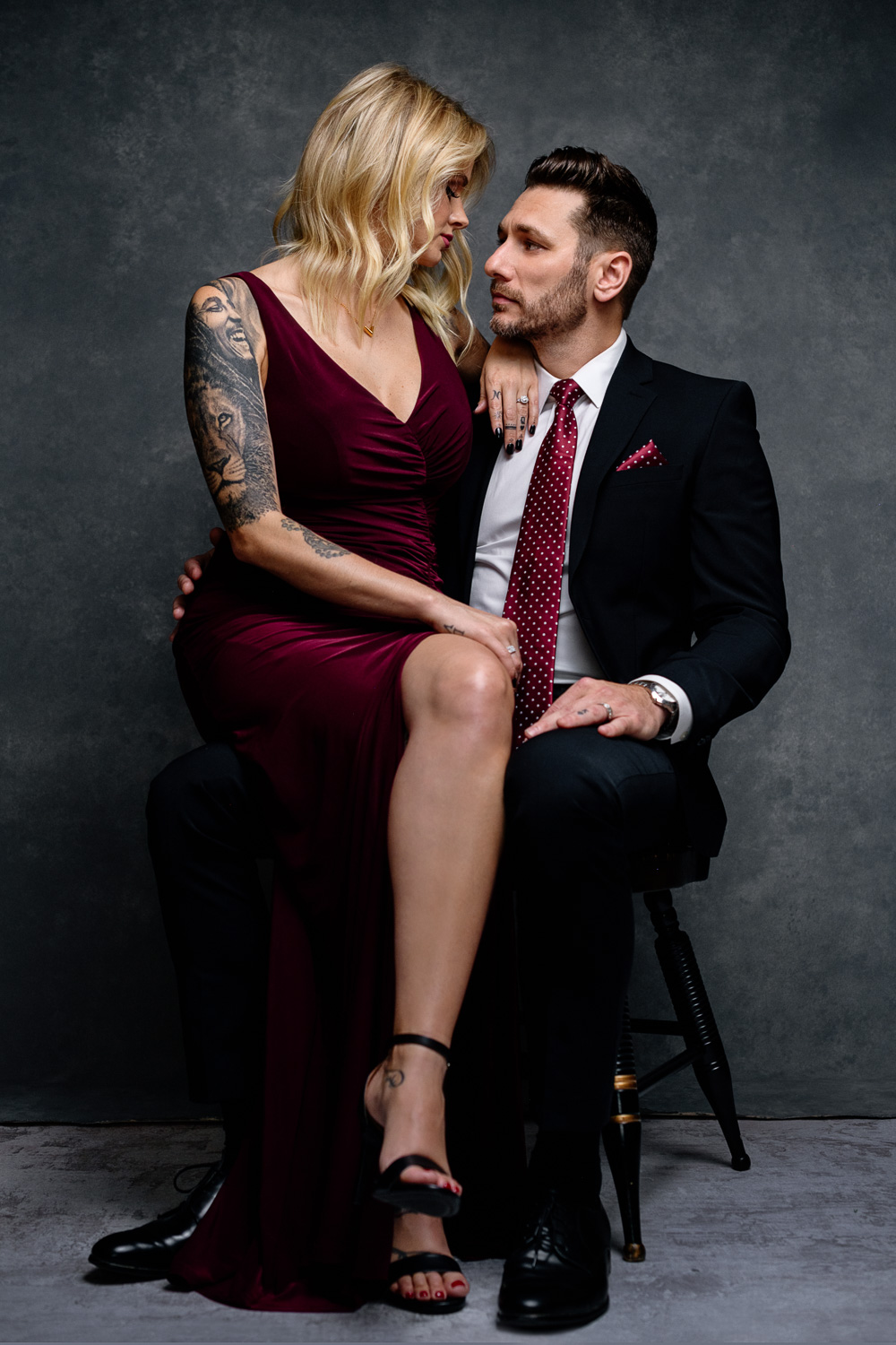 Beautiful blonde woman wearing burgundy dress with tattoos sits on husbands leg while sharing a chair