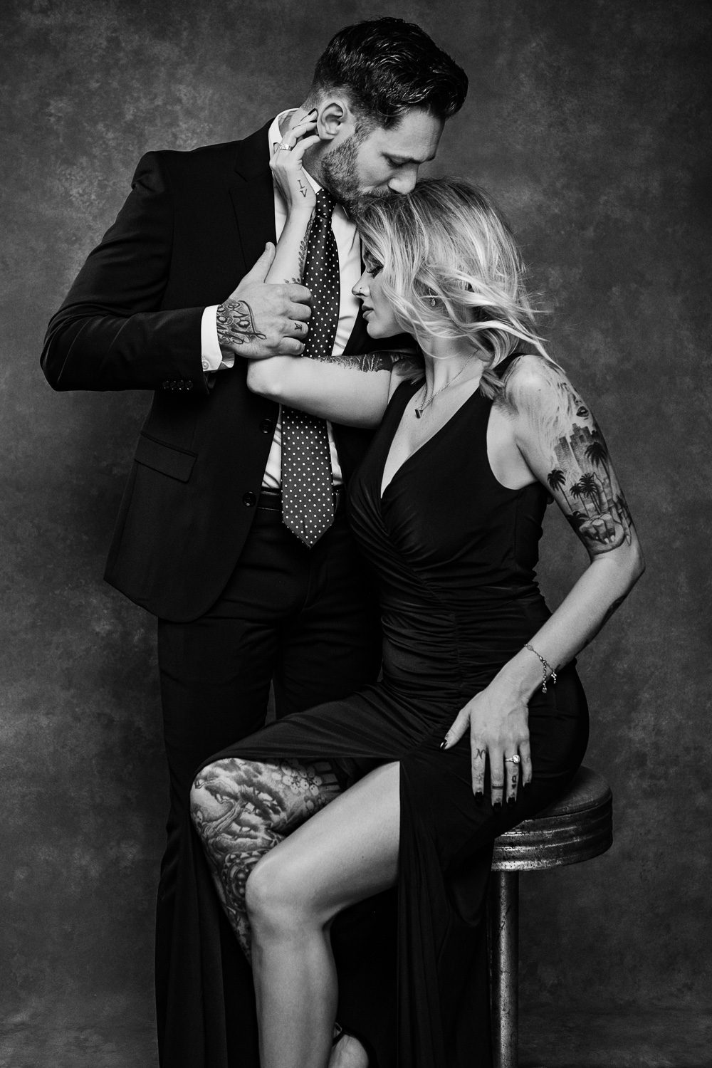 Black and white image of husband kissing wife as she sits on a vintage bar stool.