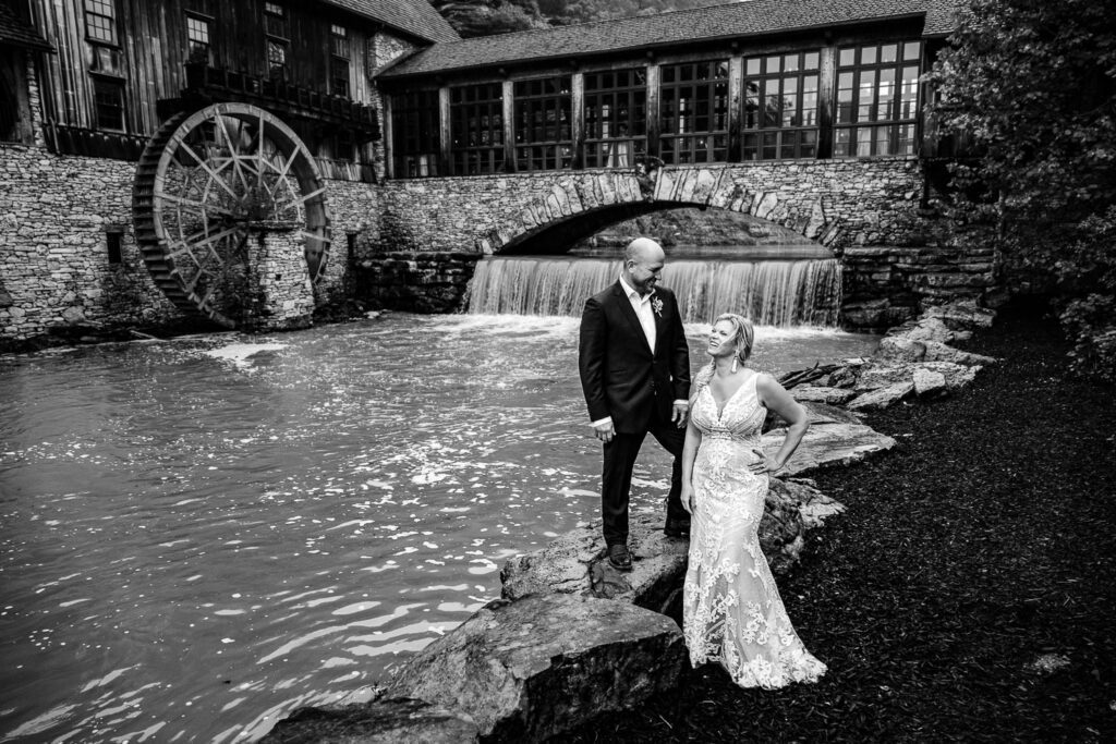 Black and white portrait of bride laughing as groom stands on rock in front of Dogwood Canyon Lodge and Grist Mill Lampe Mo Schaefer Photography