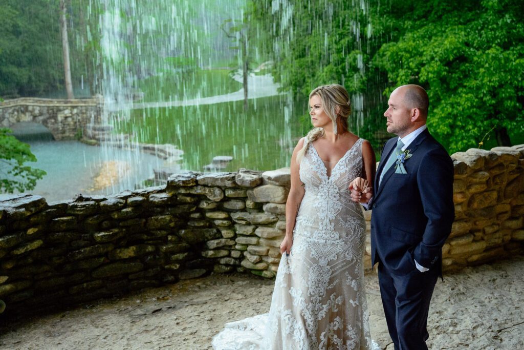 Bride and Groom hold hands as rain falls and water fall runs over rock wall