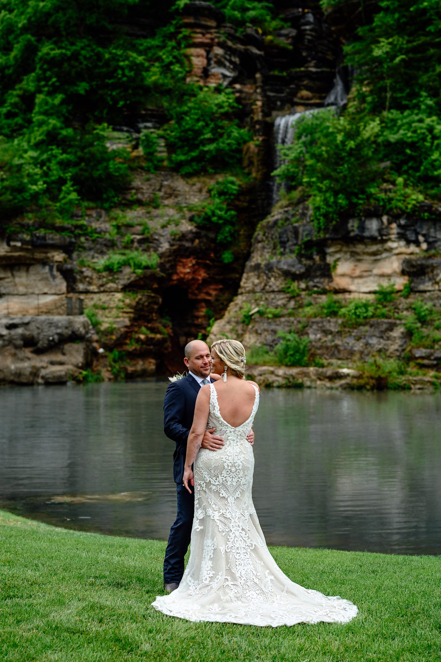 Groom holds bride's waist in front of Indian Creek and waterfall at Dogwood Canyon