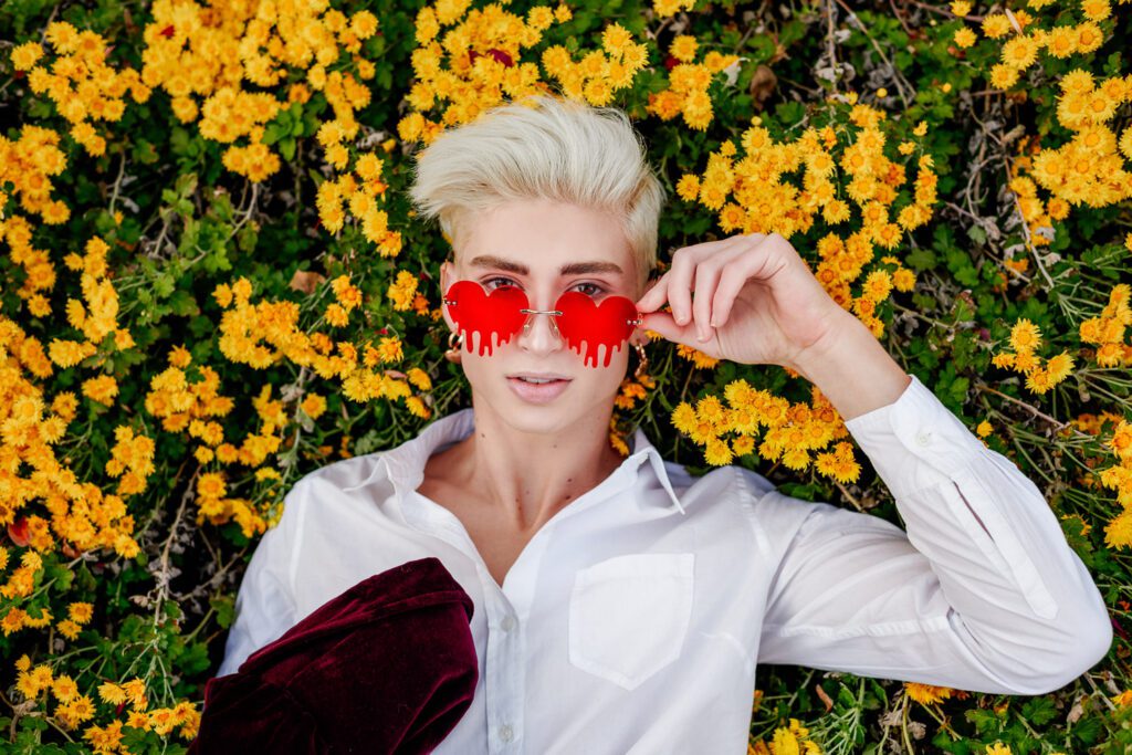 Senior guy Zachary Willmore lays in yellow flowers with red bleeding heart sunglasses. 