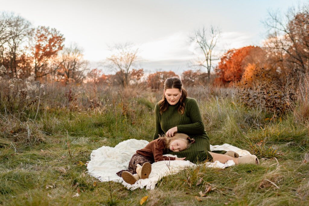 Mom and daughter sit on white blanket with fall colors.
