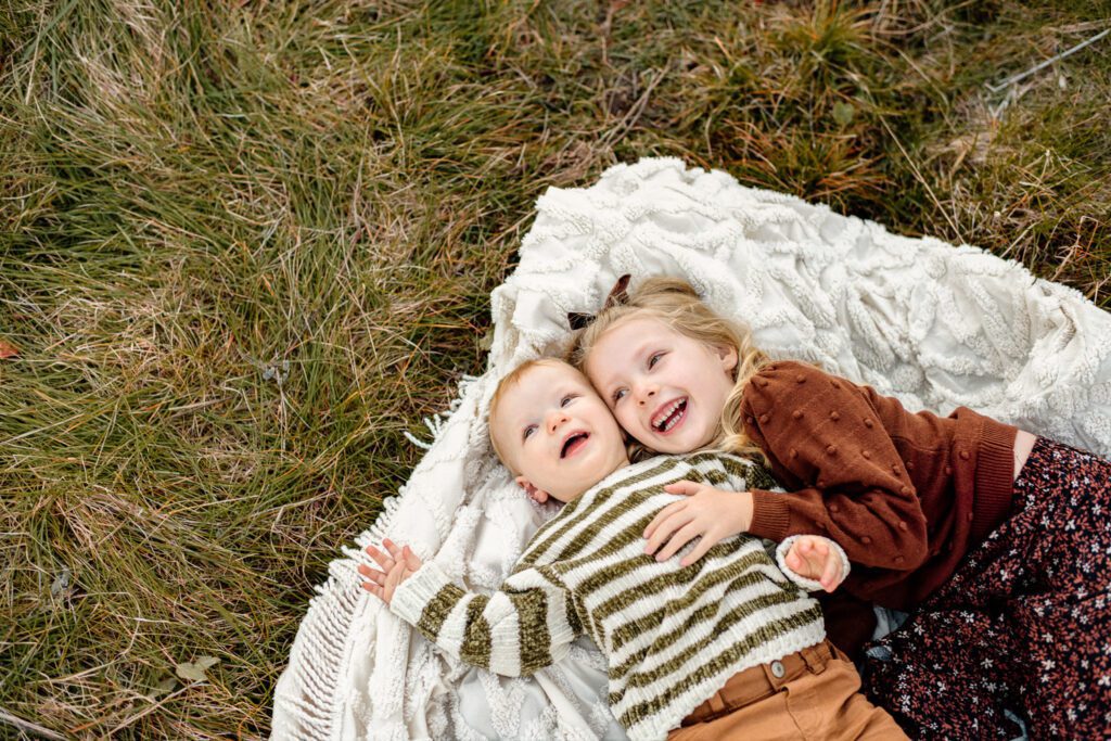 Two kids laugh on white blanket wearing green and white striped sweater and a brown sweater.