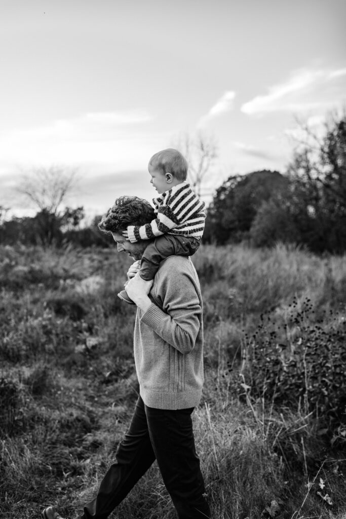 Young boy sits on father's shoulders while walking through fall field.