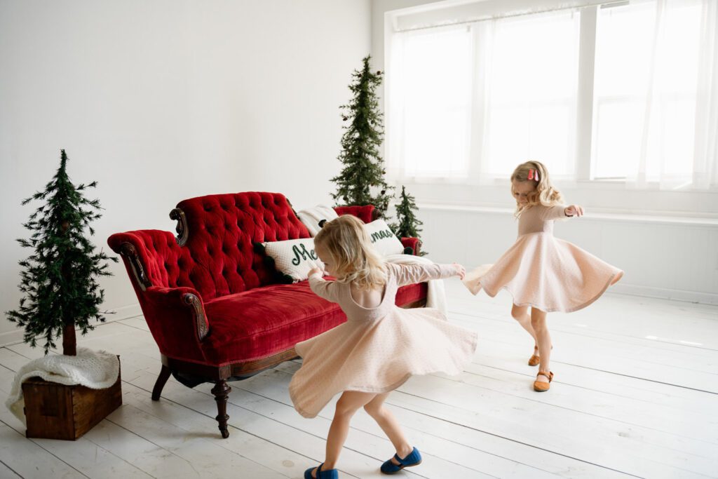 two girls dance together in front of red couch and green trees.