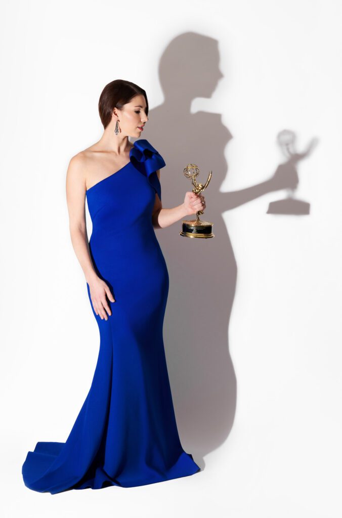 KOMU TV Anchor Emily Spain wearing a Blue Evening gown holding a NATAS Mid-America Emmy Award on a white background with shadow.