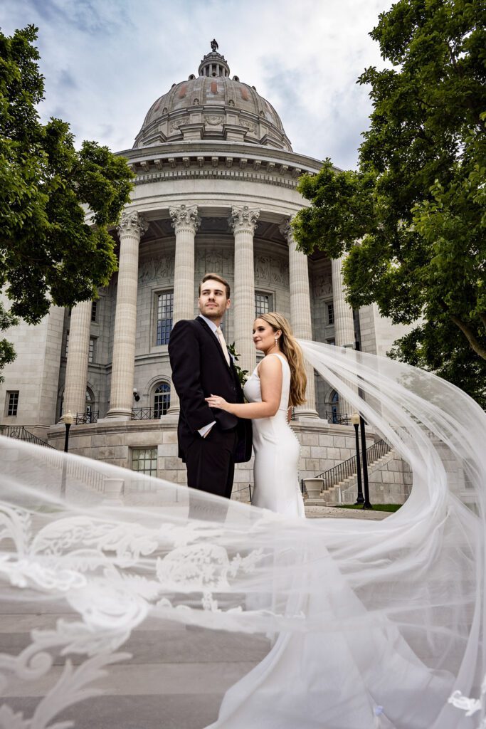 Bride and groom stand in front of majestic capitol building with veil flying in the wind.