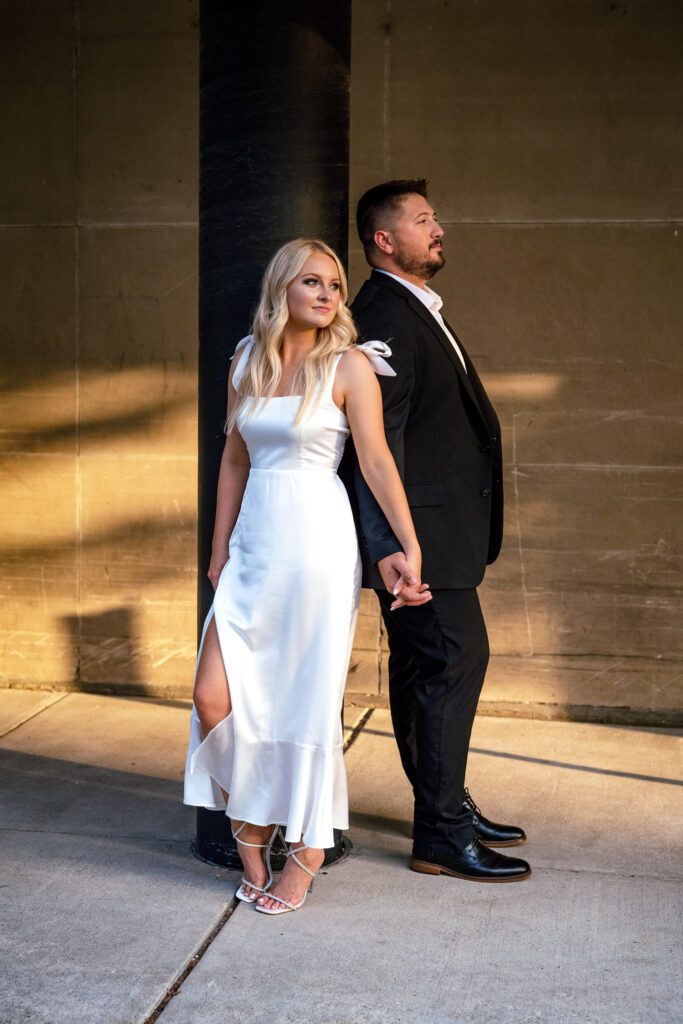 Engaged couple in black suit and white dress lean on black pole during sunset.