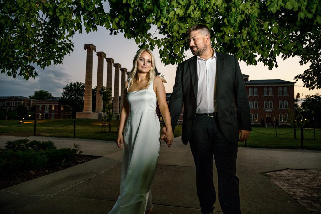 Couple walk in front of the Mizzou Columns holding hands during engagement session.