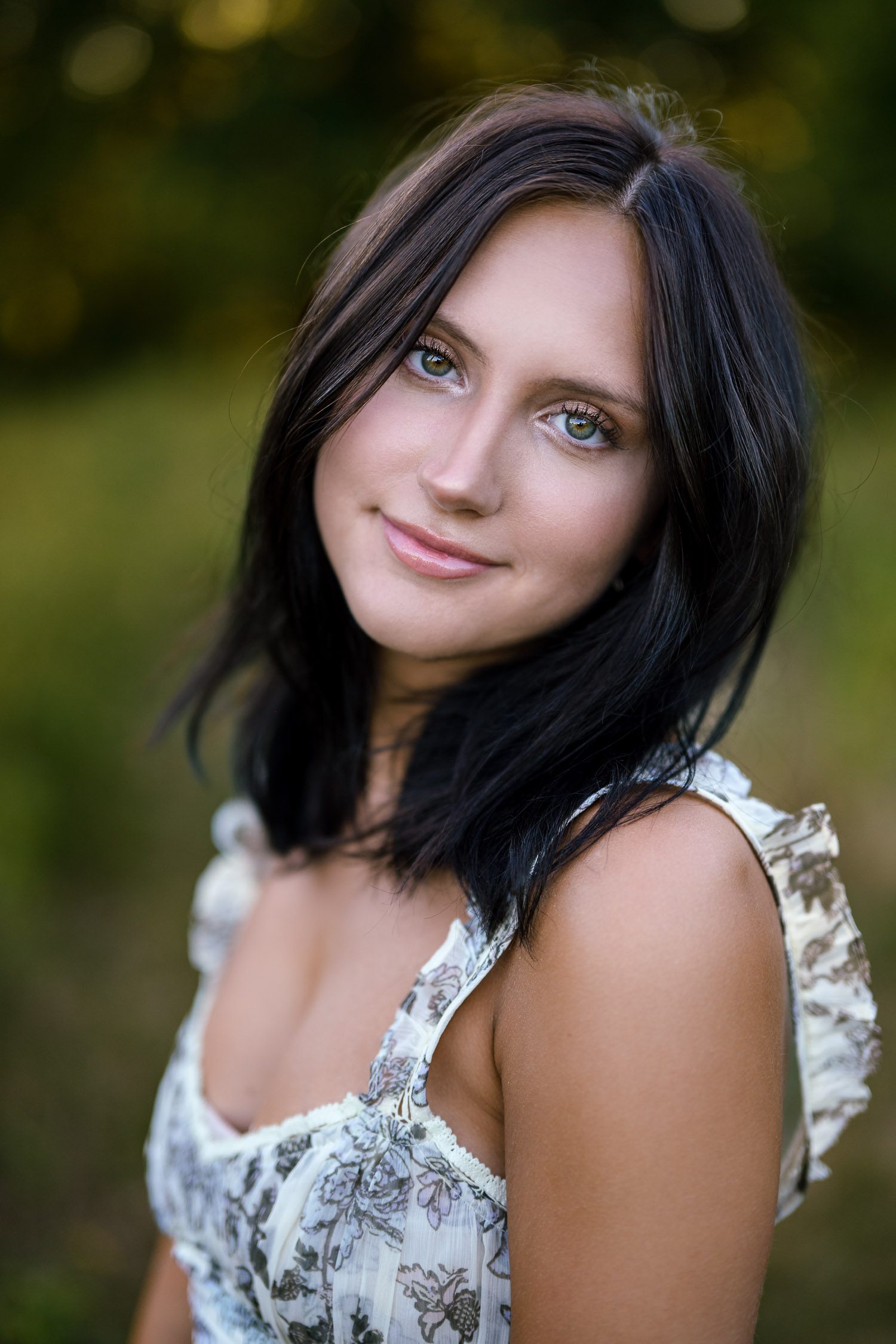 Senior girl with brunette hair and hazel eyes smiles at the camera with green nature behind her.