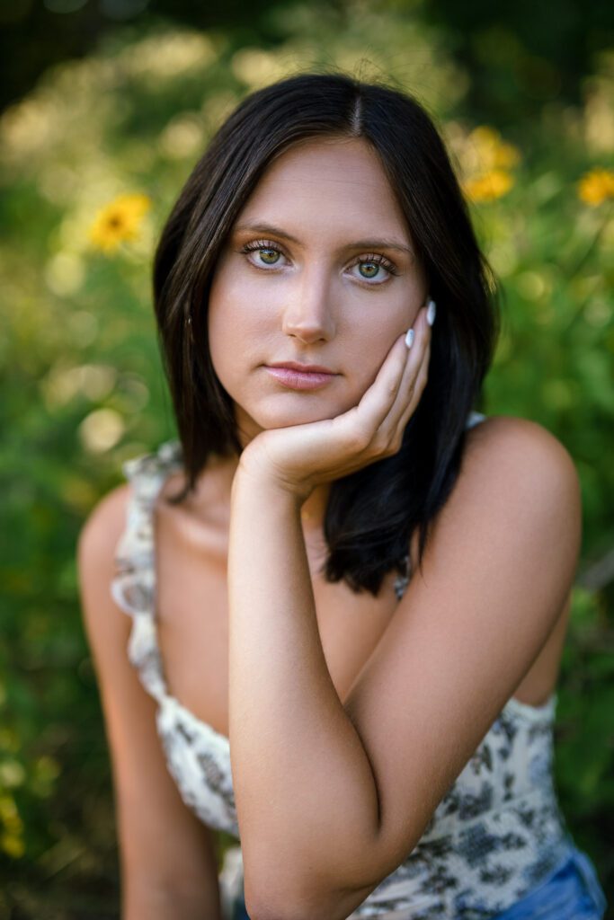 Brunette Senior girl places her chin in her hand with yellow flowers behind her.