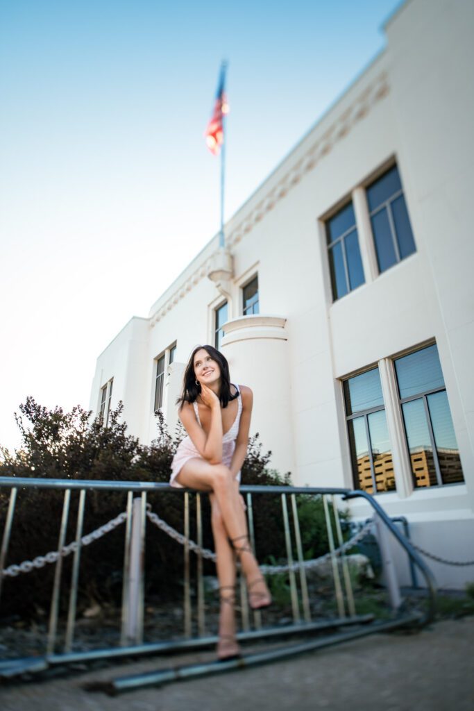 Brunette Senior girl sitting on bike rack in front of an art deco building with an american flag.