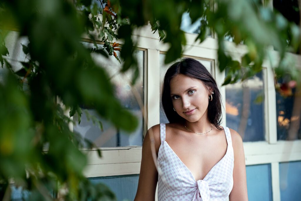 Brunette senior girl wearing a pink and white gingham romper standing in front of garage door with green leaves. 