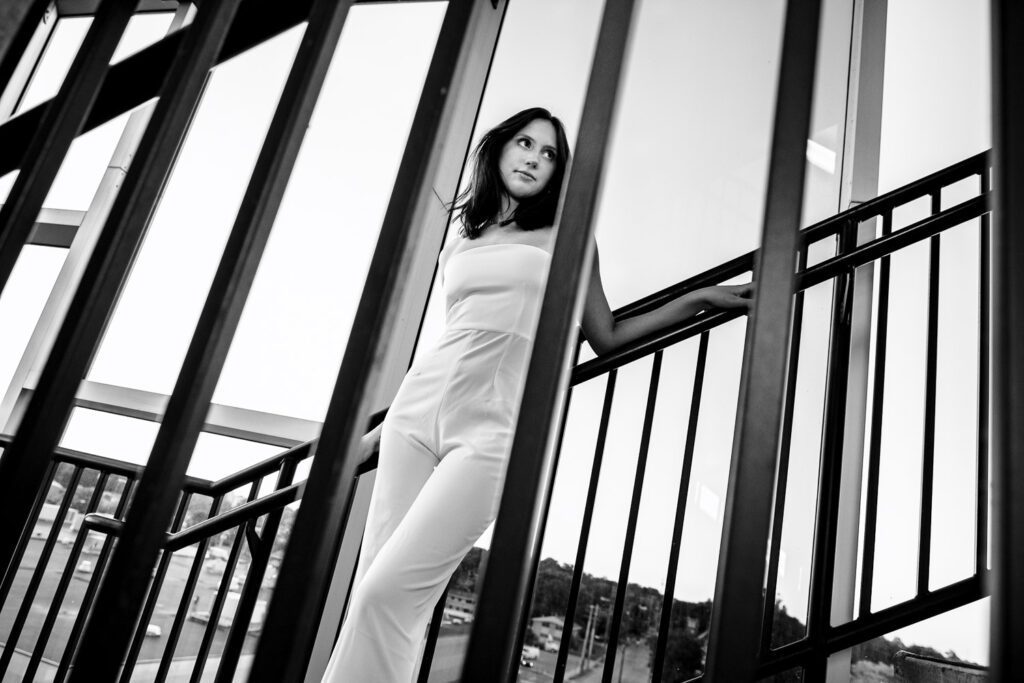 Senior girl with dark hair leans on stair railing with more railing in the foreground.