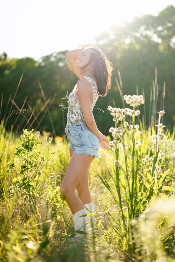 Brunette senior girl wearing white boots, jean shorts and a flower top dances in field with hand in her hair. 