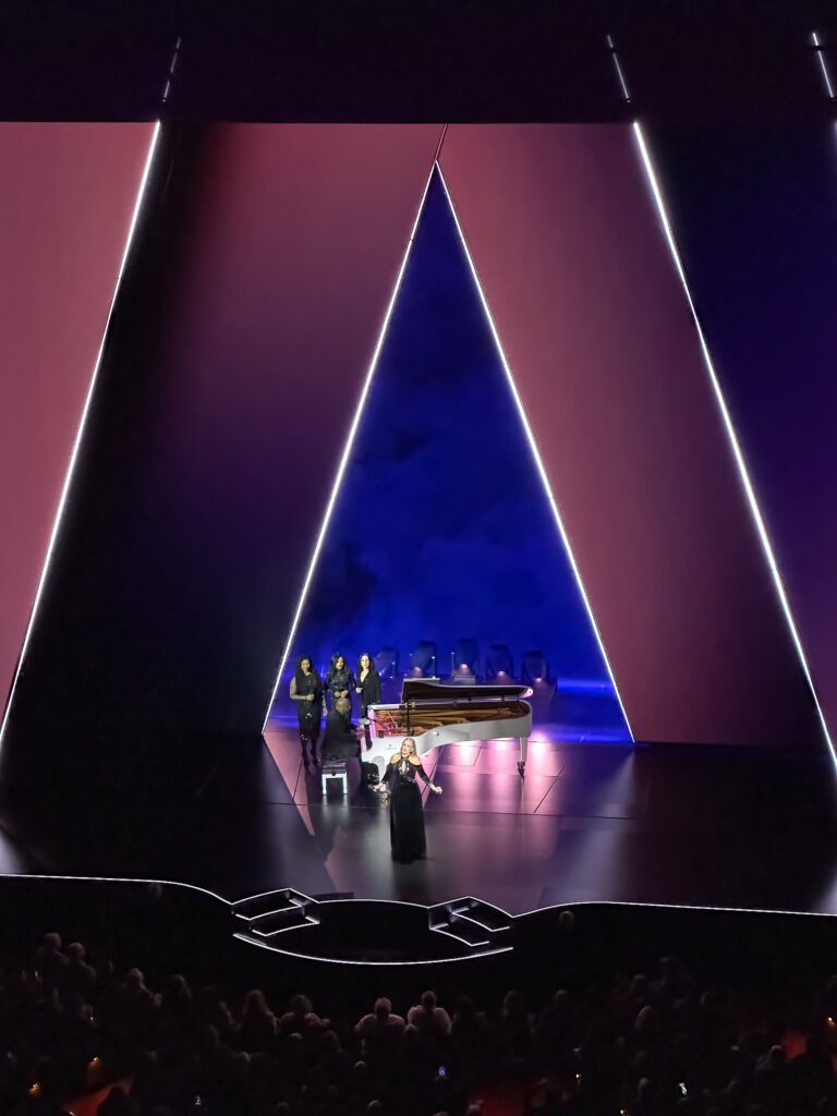 Adele sings on stage in front of A frame stage design and white piano with back up singers.