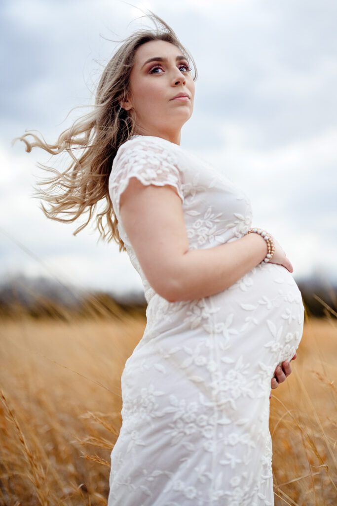 Pregnant woman wearing white dress holds baby bump in a golden field while the wind blows her hair.
