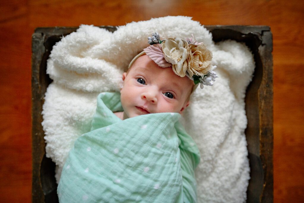 Newborn wearing flower headband looks at camera while wrapped in light green blanket.