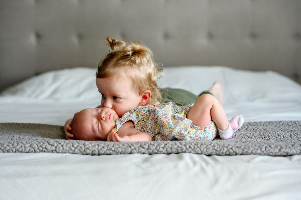 Young girl kisses newborn sister while laying on bed.