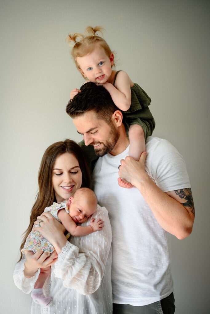 Family cuddles together with daughter on dads shoulders while looking at newborn.