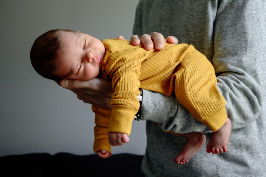 Newborn baby in yellow onesie asleep on dad's arm.