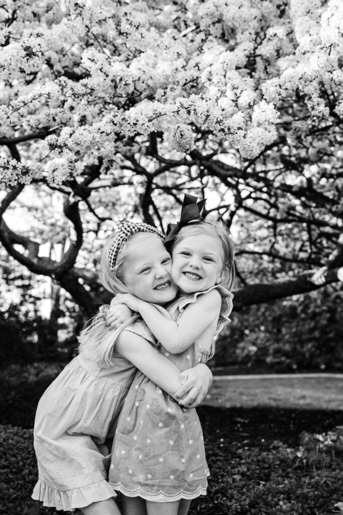 Sisters hug in front of tree with spring blossoms.