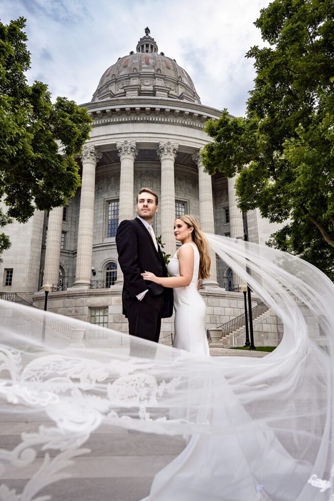 Bride and Groom pose in front of the Missouri State Capitol building in Jefferson City with long veil blowing in the wind.