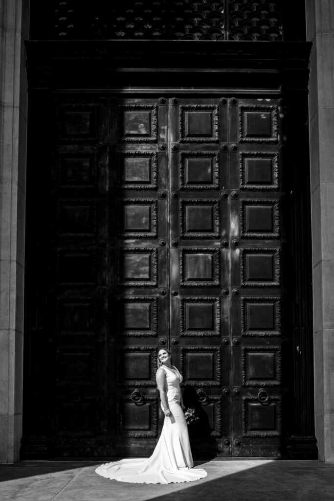 Bride stands in sunlight in front of huge black doors.