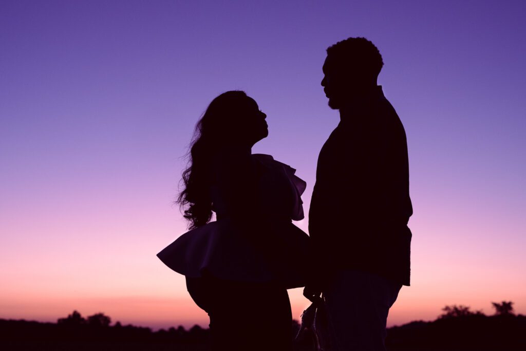 Engaged black couple look at each other with colorful sky behind their silhouette.