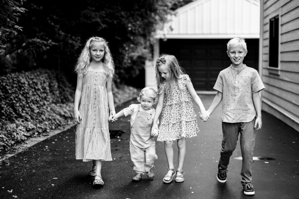Four young kids hold hand while walking down drive way.