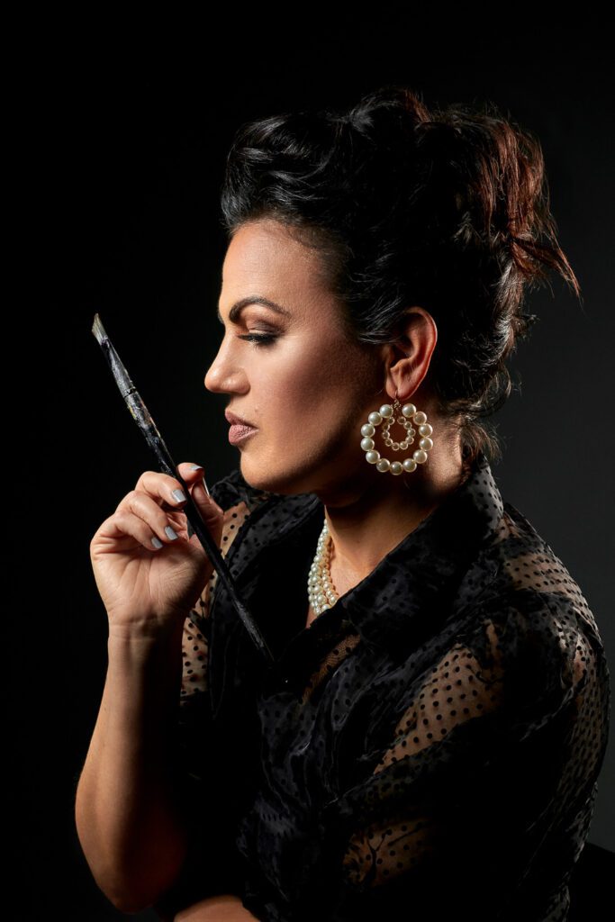 Artist Cindy Scott poses in studio with a paint brush and pearl ear rings.