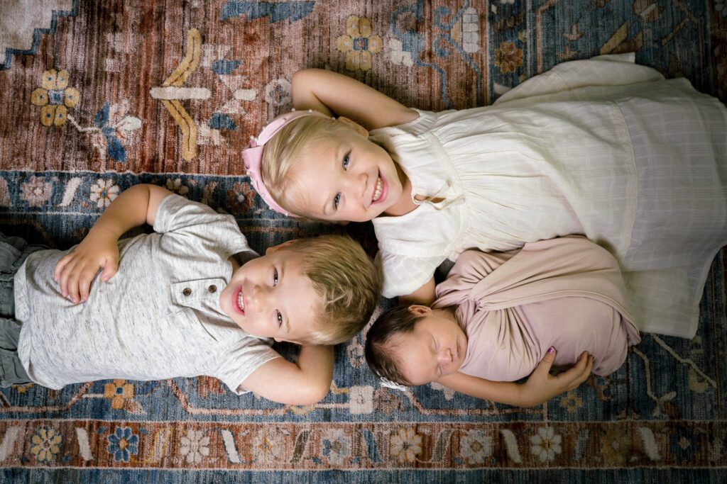 Newborn swaddled as brother and sister lay on their back on pink rug.