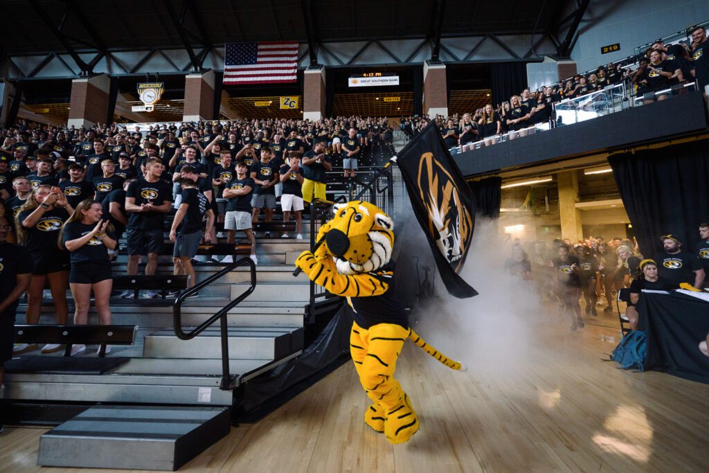 Truman the Tiger runs out of Mizzou Arena Tunnel carrying University of Missouri Flag at Student Athlete Event.