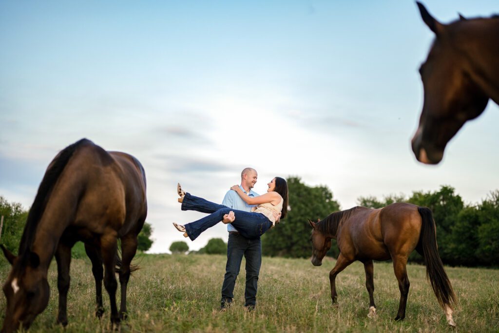 Engaged couple stand among horses in field with guy holding girl wearing cowboy boots.