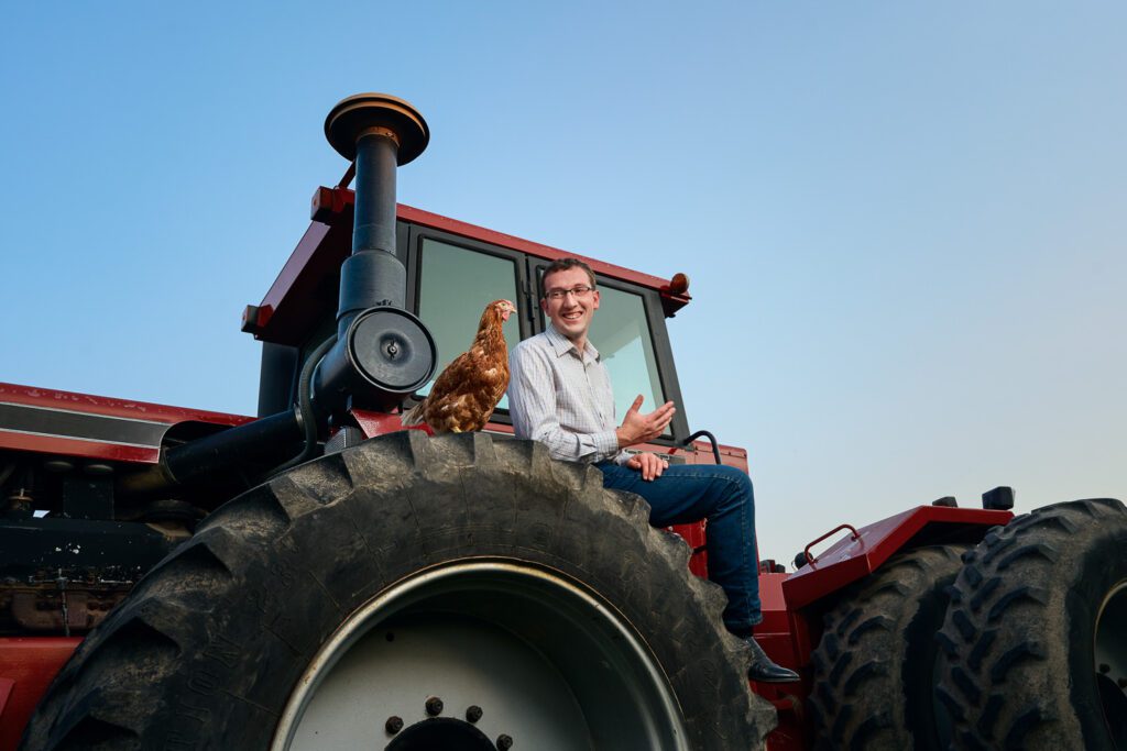 Farmer sits on top of red tractor with chicken sitting by him on wheel.