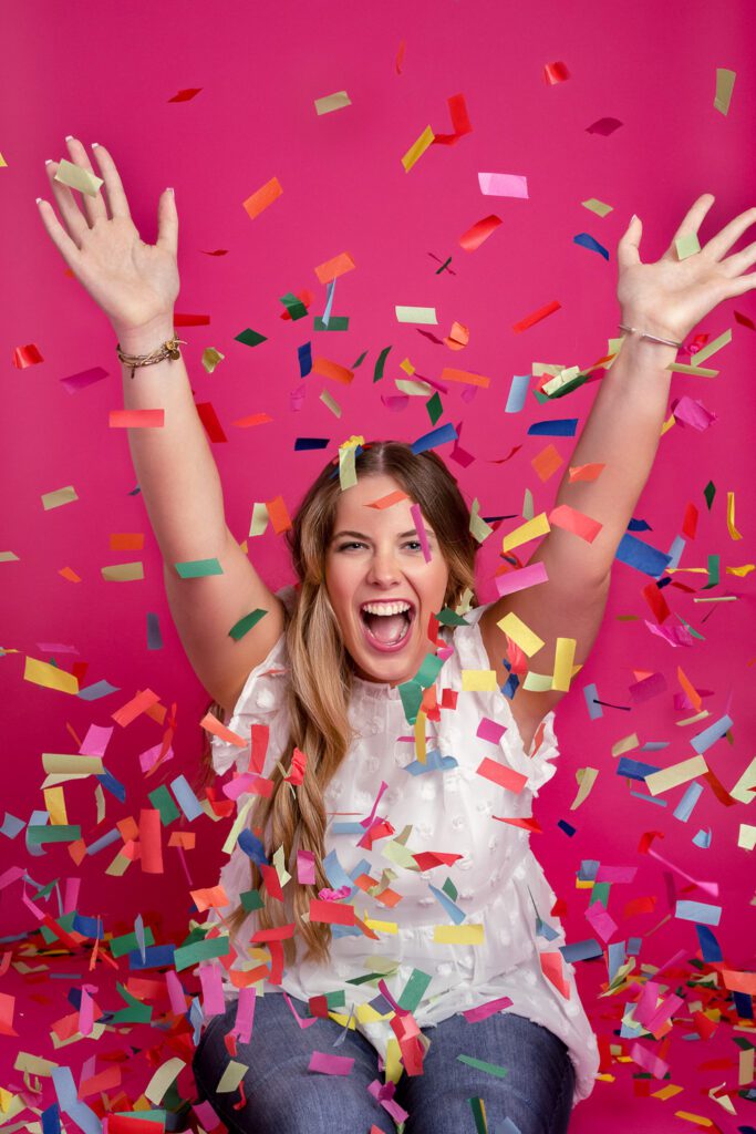 Rock Bridge Senior Girl celebrates with confetti in front of pink backdrop.